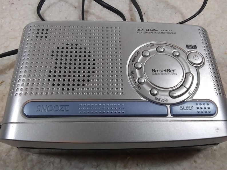 May include: Silver dual alarm clock radio with a digital display and a SmartSet auto clock set system. The radio has buttons for snooze, sleep, time zone, set, mode, and alarm.