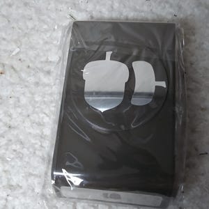 May include: A dark brown paper craft punch tool encased in clear plastic packaging. The top of the tool features a circular cutout design showing an acorn shape, and a small white label at the base displays a matching acorn icon for identification.