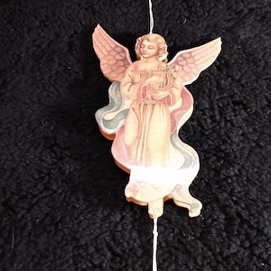May include: A wooden angel ornament with a peach and cream color scheme. The angel is holding a harp and has large wings. A white string is attached for hanging. The ornament is on a black, textured surface.