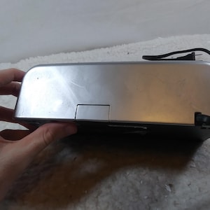May include: A silver-coloured electronic device with a black power cord. The device has a rectangular shape and a small rectangular opening on the top.