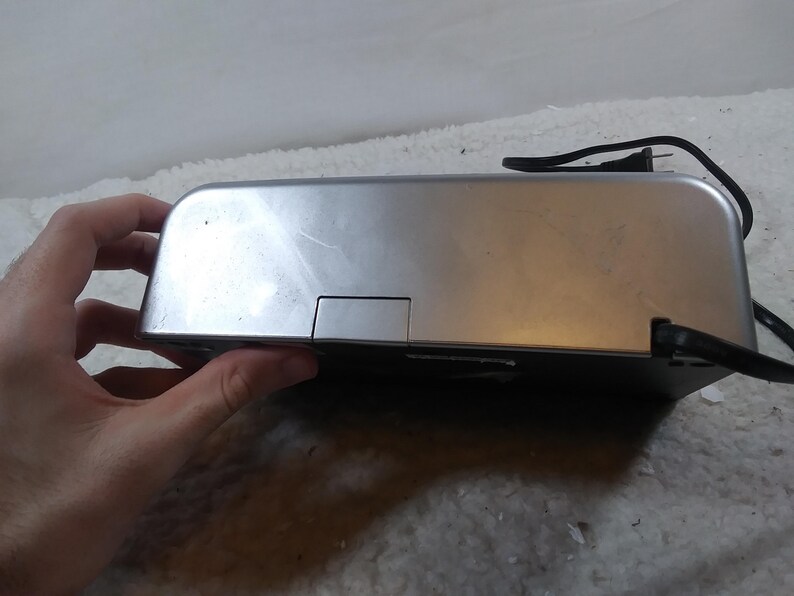 May include: A silver-coloured electronic device with a black power cord. The device has a rectangular shape and a small rectangular opening on the top.