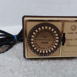 May include: A brown and gold 24-hour timer with a round dial and a black cord. The dial has numbers 1 through 12 and the words "DAY" and "NIGHT". The timer has a switch for "Outlet ON", "Outlet OFF", and "AUTOMATIC".