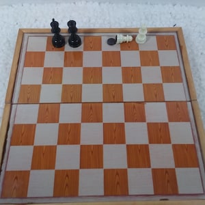 Könnte beinhalten: Ein faltbares Schachbrett aus Holz mit einem braunen und weißen Schachbrettmuster. Auf dem Brett befinden sich zwei schwarze und drei weiße Schachfiguren.