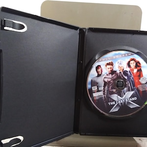 Puede incluir: Una caja de DVD negra abierta revela un disco con el título de la película "The Last Stand". El disco presenta imágenes de los personajes de la película. La caja está abierta, mostrando el disco y el interior de la caja.