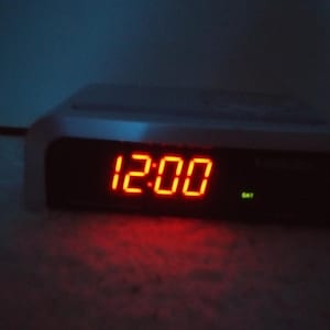 May include: A white digital clock with a red LED display showing the time 12:00.