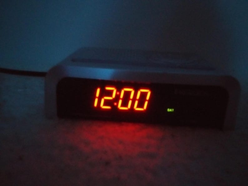 May include: A white digital clock with a red LED display showing the time 12:00.