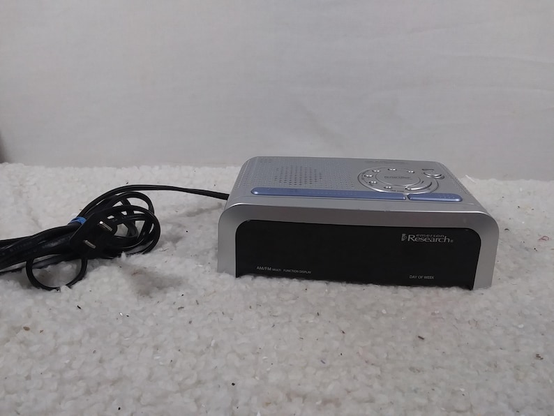 May include: A silver and black digital clock radio with a blue display and the text "Insignia by Best Buy" and "Research Plus" on the front.