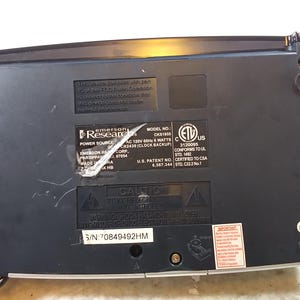 May include: Black Emerson Research clock radio with a silver face and a black and silver control panel. The back of the radio has a label with the model number CKS1855, power source information, and safety certifications. The serial number is 70849492HM.