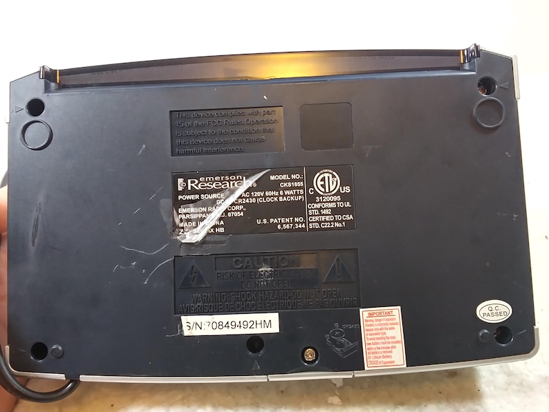 May include: Black Emerson Research clock radio with a silver face and a black and silver control panel. The back of the radio has a label with the model number CKS1855, power source information, and safety certifications. The serial number is 70849492HM.