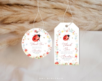 May include: Two white paper gift tags, one circular and one rectangular, featuring a watercolor ladybug, colorful wildflowers, and mushrooms. The text reads Thank You for celebrating this special day with us, Amelia's First Birthday, 06.22.2026.