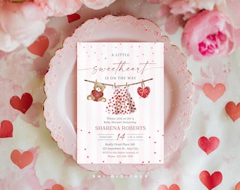 May include: A baby shower invitation with a pink and white design. The invitation features the text "A Little Sweetheart Is On The Way" and includes a teddy bear, a dress, and a heart hanging on a clothesline. The invitation is on a pink plate with red hearts.