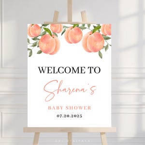 May include: A white sign on a wooden easel with the text "WELCOME TO Sharena's BABY SHOWER 07.20.2025". The top of the sign features a watercolour design of peaches and green leaves.