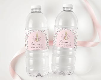 May include: Two clear plastic water bottles with white caps. Each bottle has a pink label with a bunny illustration and the text "Sharena's Baby Shower 01.20.2025". Floral accents surround the bunny. A pink ribbon is in the background.