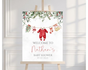 May include: A welcome sign for Nathan's baby shower, featuring a watercolor design with Christmas-themed elements. The sign includes a red onesie, stocking, and bell hanging on a string, with greenery and the date 09.05.2025.