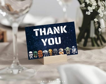 以下が含まれることがあります： 紺色の「Thank You」カード。白い文字と星空の背景に、スターウォーズのキャラクターフィギュアが描かれています。カードは木製のブロックに立てられており、パーティーやイベントに適しています。