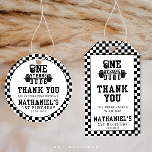 May include: Two gift tags with a fitness theme. One is round, the other rectangular. Both have a black and white checkered border and text that reads "ONE STRONG DUDE THANK YOU" and "NATHANIEL'S 1ST BIRTHDAY 02.04.2026".