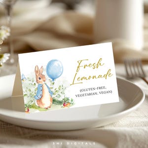 May include: A white card with the text "Fresh Lemonade" and a watercolor illustration of a bunny holding a blue balloon. The card also states "(Gluten-Free, Vegetarian, Vegan)". The card is on a white plate.