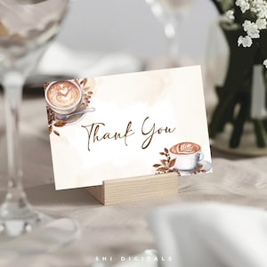 May include: A cream-colored card with the words "Thank You" in elegant script. The card features watercolor illustrations of coffee cups and coffee beans. The card is displayed in a wooden holder, perfect for a coffee-themed event.