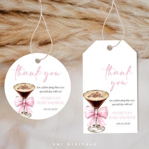 May include: Two white gift tags with a pink watercolor design. Each tag features a cocktail illustration with a pink bow, the words "thank you," and text for a baby shower. The tags are connected to a string.