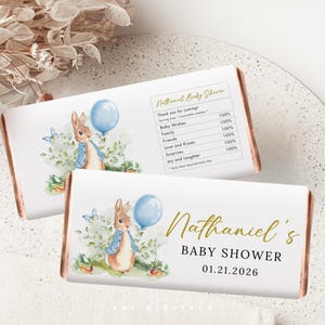 May include: Two baby shower candy bar wrappers. Each wrapper features a watercolor illustration of a bunny holding a blue balloon. The text "Nathaniel's BABY SHOWER 01.21.2026" is displayed in gold lettering.