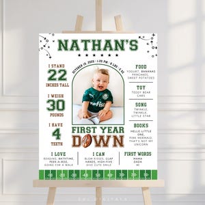 May include: A personalized first birthday sign on an easel. The sign features a photo of a baby, with text including the baby's name, height (22 inches), weight (30 pounds), and other details. The sign has a football theme.