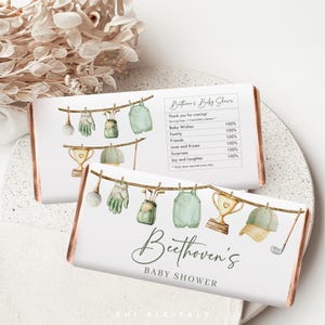 Pode incluir: Duas embalagens brancas de barras de chocolate com um design de chá de bebé com tema de golfe. As embalagens apresentam ilustrações em aquarela de equipamentos de golfe e o texto "Beethoven's BABY SHOWER". O design inclui um varal com bolas de golfe, luvas e um troféu.
