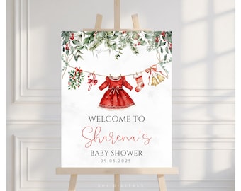 May include: A watercolor baby shower sign on an easel. The sign features a red dress, stocking, and bells hanging from a string, with greenery at the top. Text reads "Welcome to Sharena's Baby Shower 09.05.2025."