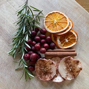 Puede incluir: Rodajas de naranja secas, rodajas de manzana, arándanos rojos y ramitas de romero dispuestas sobre una superficie de madera. Las rodajas de manzana están espolvoreadas con canela.