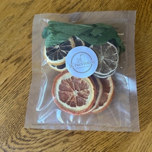 May include: A clear plastic bag filled with dried orange slices, mint leaves, and dried lemon slices. A circular sticker with the text "JK Provisions Homemade Goodness" is visible. The bag is sealed and sits on a wooden surface.