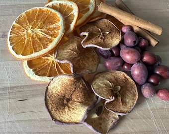 May include: A collection of dried orange slices, dehydrated apple rings dusted with cinnamon, two whole cinnamon sticks, and a cluster of small, dark purple berries arranged together. The items are grouped on a light-colored wooden surface.