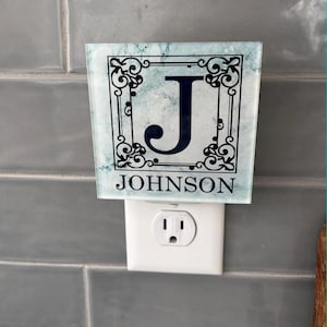 May include: A decorative night light with a marble-effect background. The night light features a black ornate border, a large black letter "J", and the name "JOHNSON" in black text. The night light is plugged into a white electrical outlet.