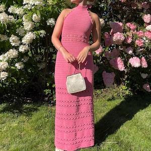 Puede incluir: Vestido halter rosa de ganchillo con cuello alto y corpiño ajustado. El vestido es largo hasta el suelo y presenta un patrón texturizado. Se sostiene un pequeño bolso plateado delante del vestido. El fondo incluye un cielo azul vibrante y un exuberante jardín con flores blancas y rosas.