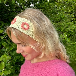 May include: A crocheted headband in a light yellow color, adorned with two pink flower motifs. The headband is worn on a person with blonde hair, against a backdrop of green foliage. The headband is a stylish accessory.