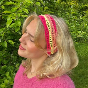 May include: A vibrant, handmade crochet headband in shades of pink, yellow, and orange. The headband features a textured design and is worn on the head. The background is a lush green with foliage.