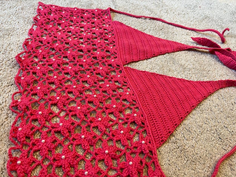 May include: A red crocheted halter top with a delicate floral pattern and white beads. The top has a V-shaped neckline and adjustable straps.