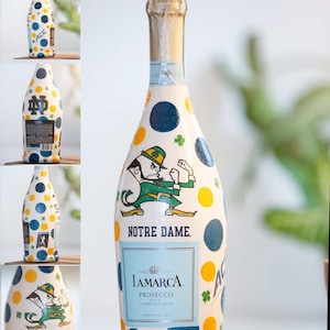 May include: A bottle of Iamarca Prosecco sparkling wine, hand-painted with a white base and colorful polka dots. The bottle features a Notre Dame Fighting Irish mascot illustration, shamrocks, and the text "Notre Dame."