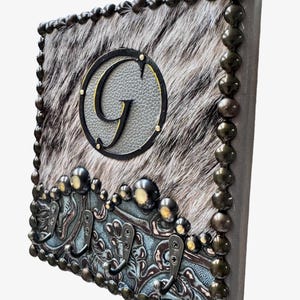 May include: Square wall decor featuring a cowhide pattern with a large letter "G" in a circle. The border is adorned with decorative metal studs and hooks, creating a rustic, western-inspired design.