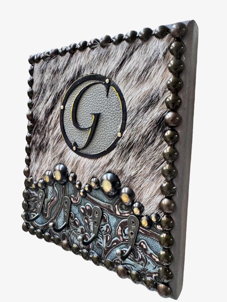 May include: Square wall decor featuring a cowhide pattern with a large letter "G" in a circle. The border is adorned with decorative metal studs and hooks, creating a rustic, western-inspired design.