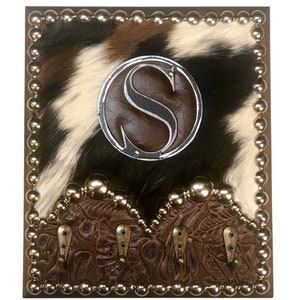 May include: A square wall-mounted coat rack with a cowhide background in black, white, and brown. It features a brown leather border with silver studs, a circular emblem with the letter "S", and four metal hooks.