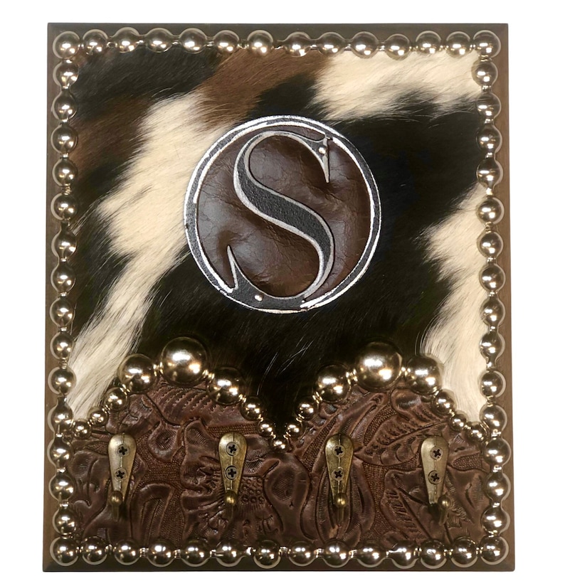 May include: A square wall-mounted coat rack with a cowhide background in black, white, and brown. It features a brown leather border with silver studs, a circular emblem with the letter "S", and four metal hooks.