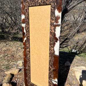 May include: A rectangular mirror with a brown and white cowhide frame. The frame is adorned with a decorative embossed pattern and small silver studs. The mirror has a light brown backing.