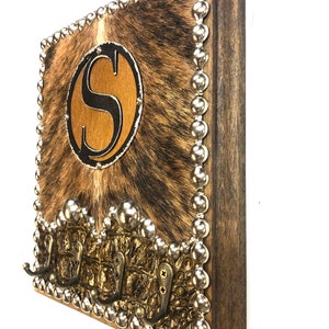 May include: A rustic wooden wall-mounted key holder with a cowhide-like center and a large letter "S" in a circular design. The border is adorned with silver studs, and it features three brass hooks for keys or other items.