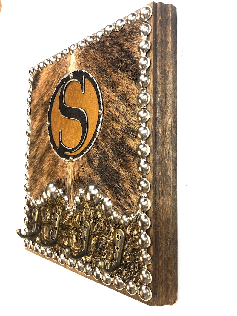 May include: A rustic wooden wall-mounted key holder with a cowhide-like center and a large letter "S" in a circular design. The border is adorned with silver studs, and it features three brass hooks for keys or other items.