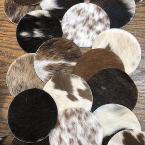 May include: A collection of round coasters made from cowhide. The coasters display a variety of natural patterns and colors, including shades of brown, black, and white. The cowhide texture is visible, adding a rustic and unique touch to each coaster.