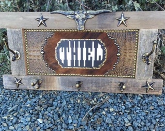 May include: Rustic wooden wall-mounted coat rack with a Western theme. Features a central panel with the word "WHITE" in a stylized font, surrounded by faux leather and metal studs. Includes hooks, star accents, and a steer head.