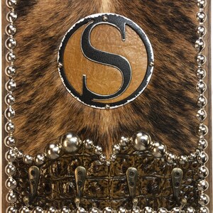 May include: A square wooden wall hook with a cowhide background and a brown faux alligator skin bottom. The center features a metal circle with the letter "S". Silver ball studs border the edges and the faux alligator skin.