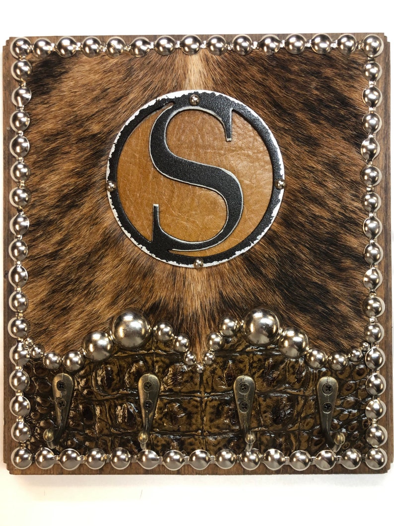 May include: A square wooden wall hook with a cowhide background and a brown faux alligator skin bottom. The center features a metal circle with the letter "S". Silver ball studs border the edges and the faux alligator skin.