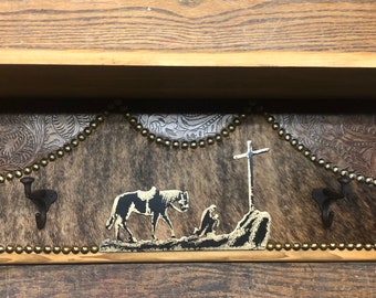 May include: Wooden shelf with a cowhide-style backing, adorned with a cross and a silhouette of a horse and rider. Features decorative metal hooks and a beaded border. The shelf is approximately 24 inches wide.