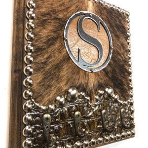 May include: Rustic key holder with a cowhide center, a brown leather-like circle with the letter "S", and a metal border. The bottom features a brown, crocodile-textured section with three metal hooks. The edges are trimmed with silver studs.