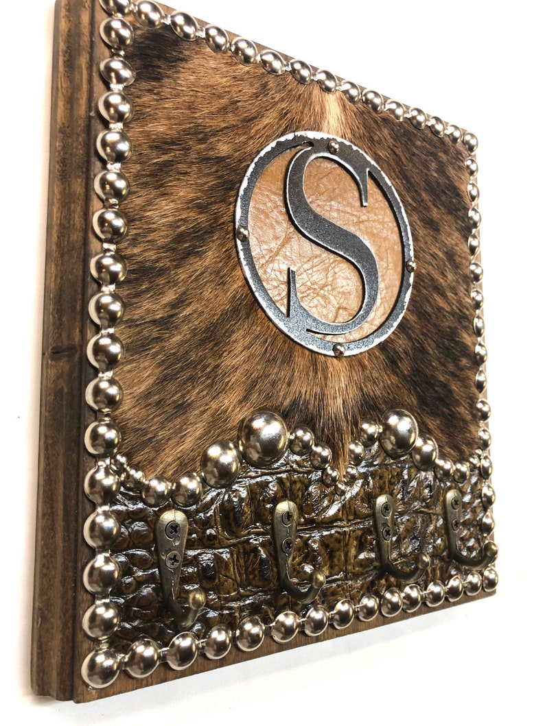 May include: Rustic key holder with a cowhide center, a brown leather-like circle with the letter "S", and a metal border. The bottom features a brown, crocodile-textured section with three metal hooks. The edges are trimmed with silver studs.